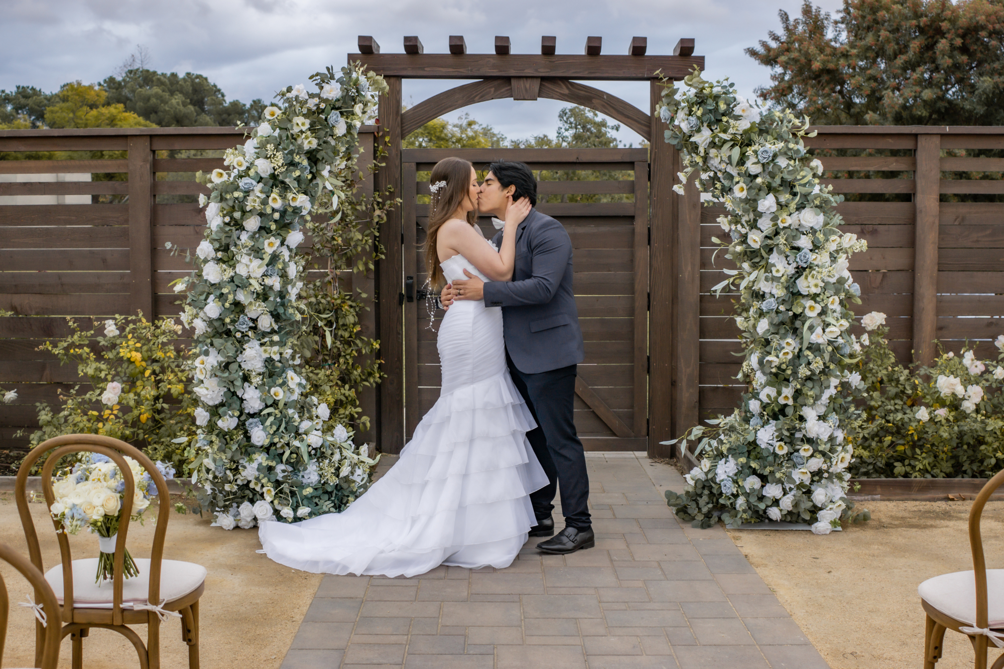 Bride and groom sharing a kiss during an outdoor garden wedding ceremony beneath a floral arch, photographed by Fonseca Hansen Media.